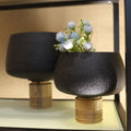 Two black ceramic planters with gold bases on a reflective surface.