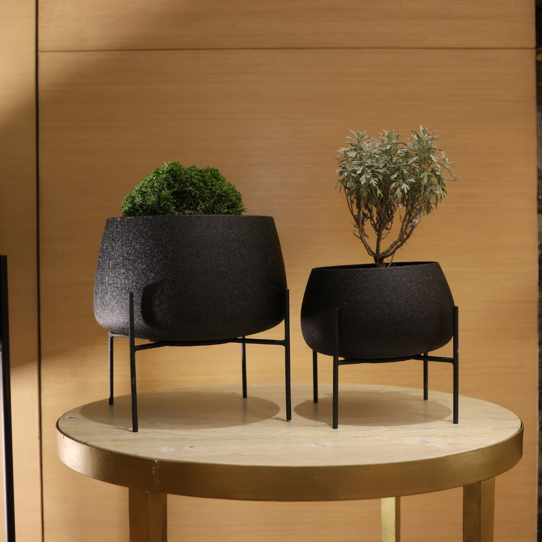 Two black textured planters with plants on a round wooden table against a wooden wall.